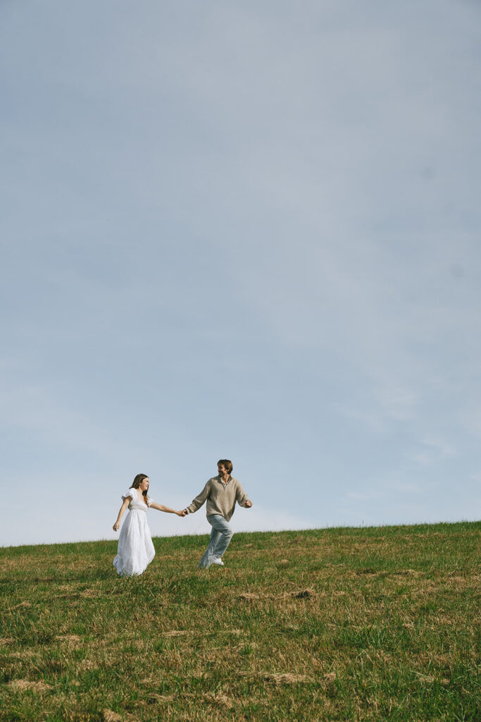 A couple holding hands walks up a grassy hill under a clear sky | What to Wear for Engagement Photos