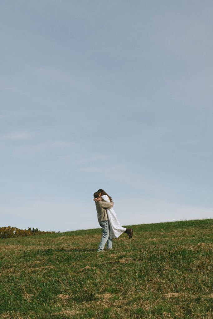 A couple holding hands walks up a grassy hill under a clear sky | What to Wear for Engagement Photos