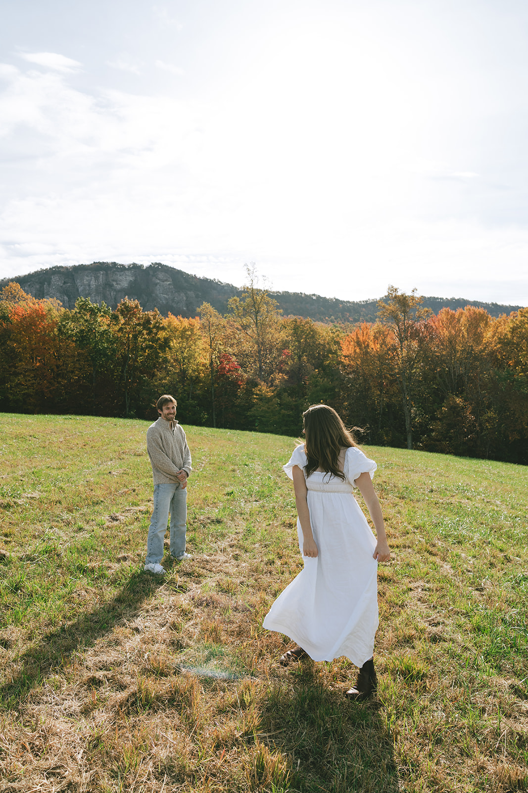 A woman in a white dress walks toward a man standing in a grassy field with trees and hills in the background under a bright sky.