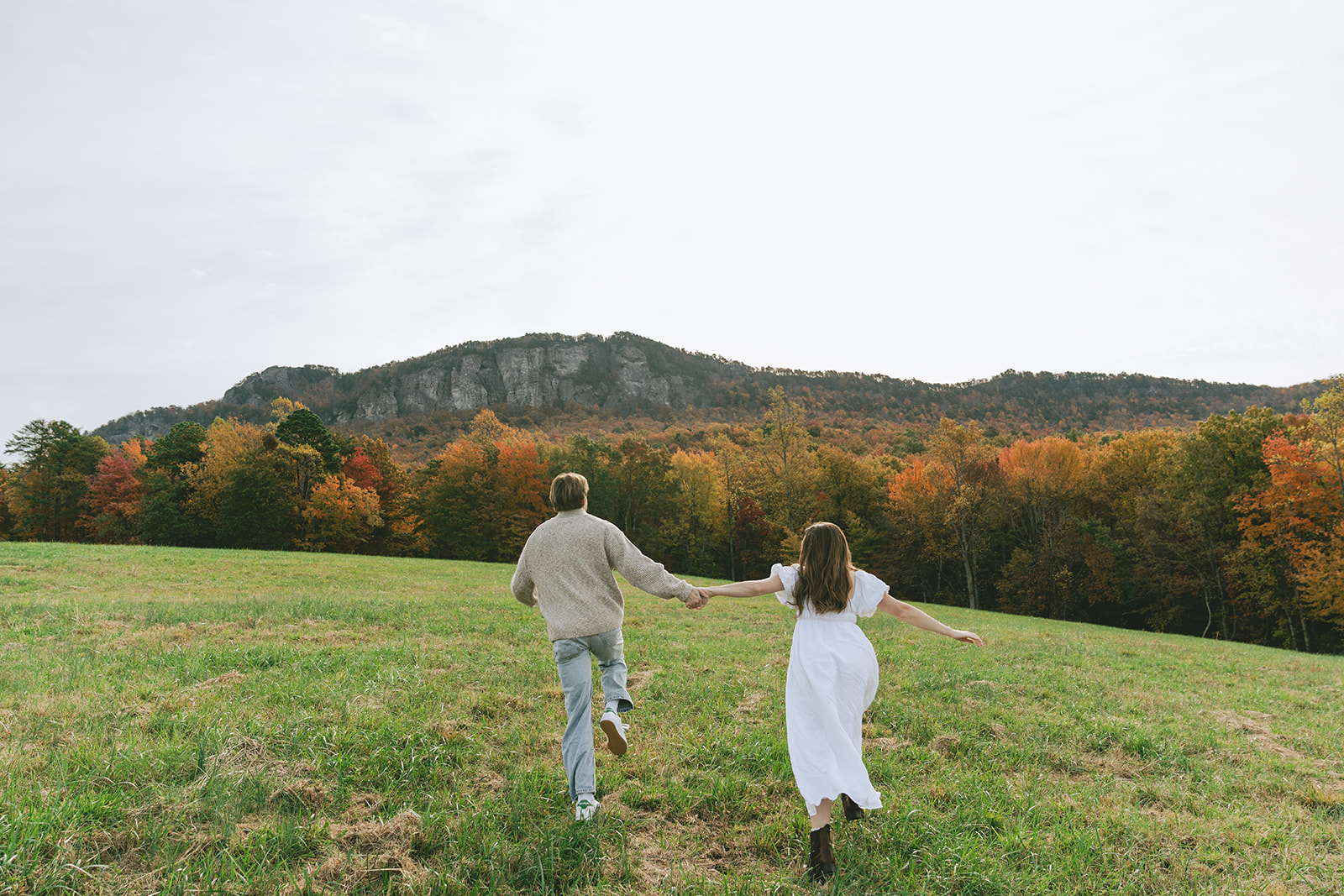 A woman in a white dress walks toward a man standing in a grassy field with trees and hills in the background under a bright sky.