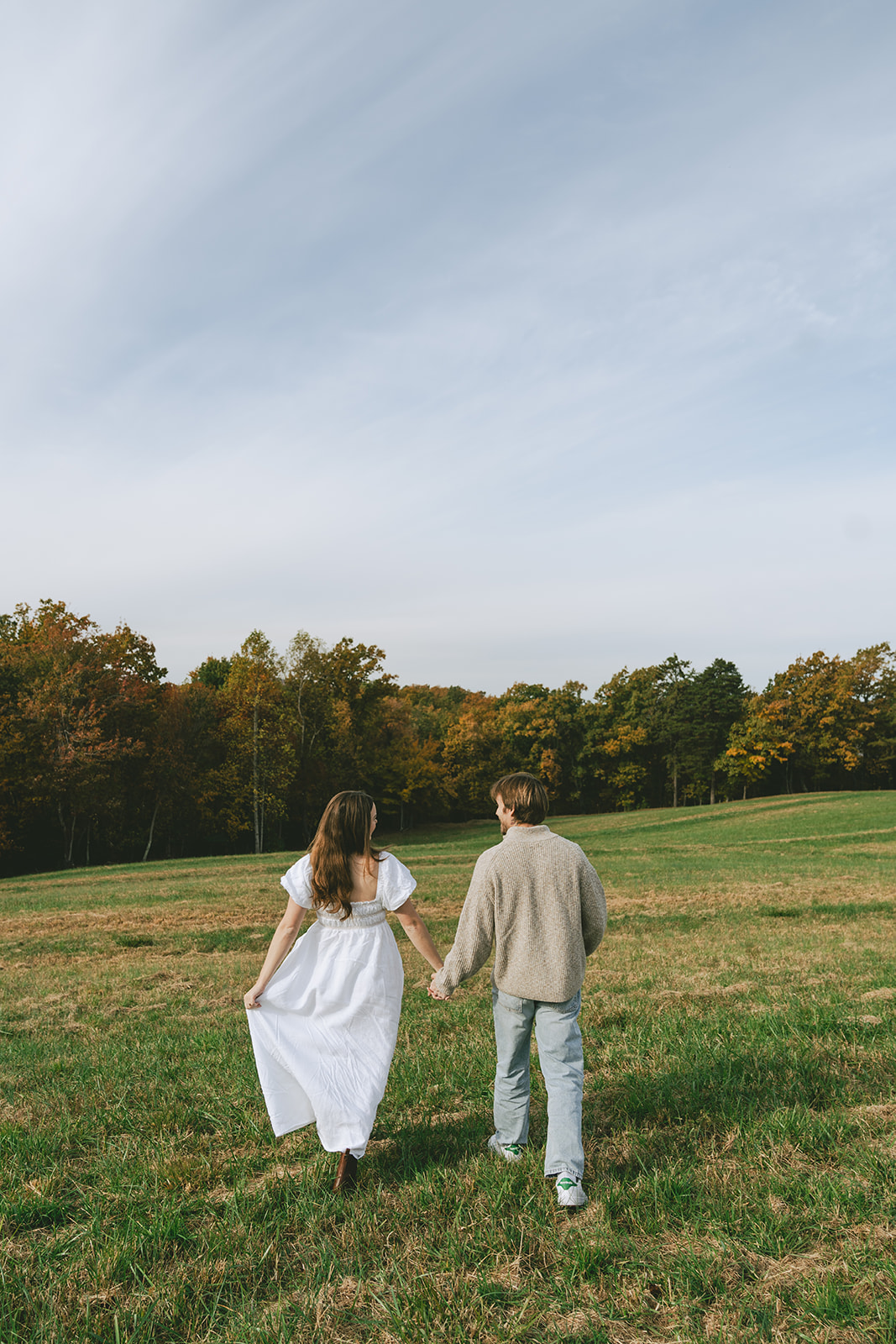 A woman in a white dress walks toward a man standing in a grassy field with trees and hills in the background under a bright sky.