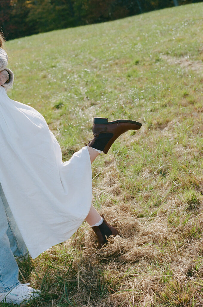 A person wearing a white dress and black ankle boots lifts their leg while standing in a grassy field.