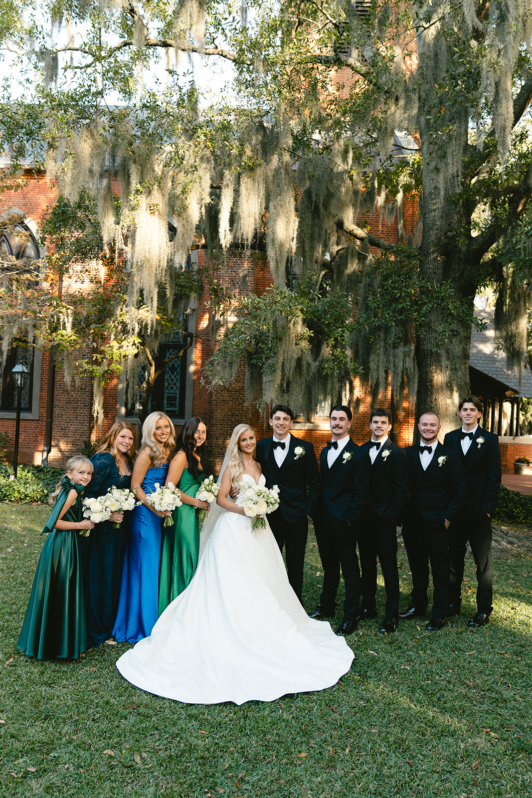 A bride in a white gown stands with her wedding party, including bridesmaids in green and blue dresses and groomsmen in black suits, outdoors near a brick building with moss-draped trees for a wedding at Stanly Hall Ballroom