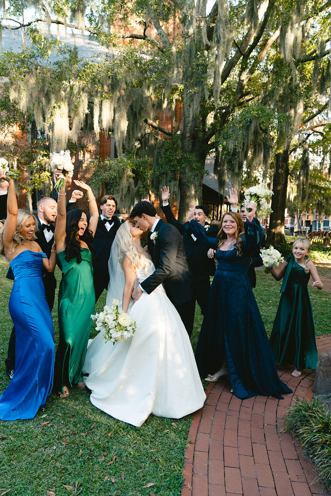 A bride in a white gown stands with her wedding party, including bridesmaids in green and blue dresses and groomsmen in black suits, outdoors near a brick building with moss-draped trees for a wedding at Stanly Hall Ballroom