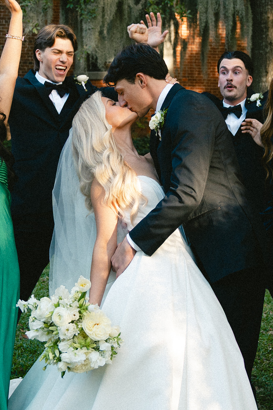 A bride in a white gown stands with her wedding party, including bridesmaids in green and blue dresses and groomsmen in black suits, outdoors near a brick building with moss-draped trees for a wedding at Stanly Hall Ballroom
