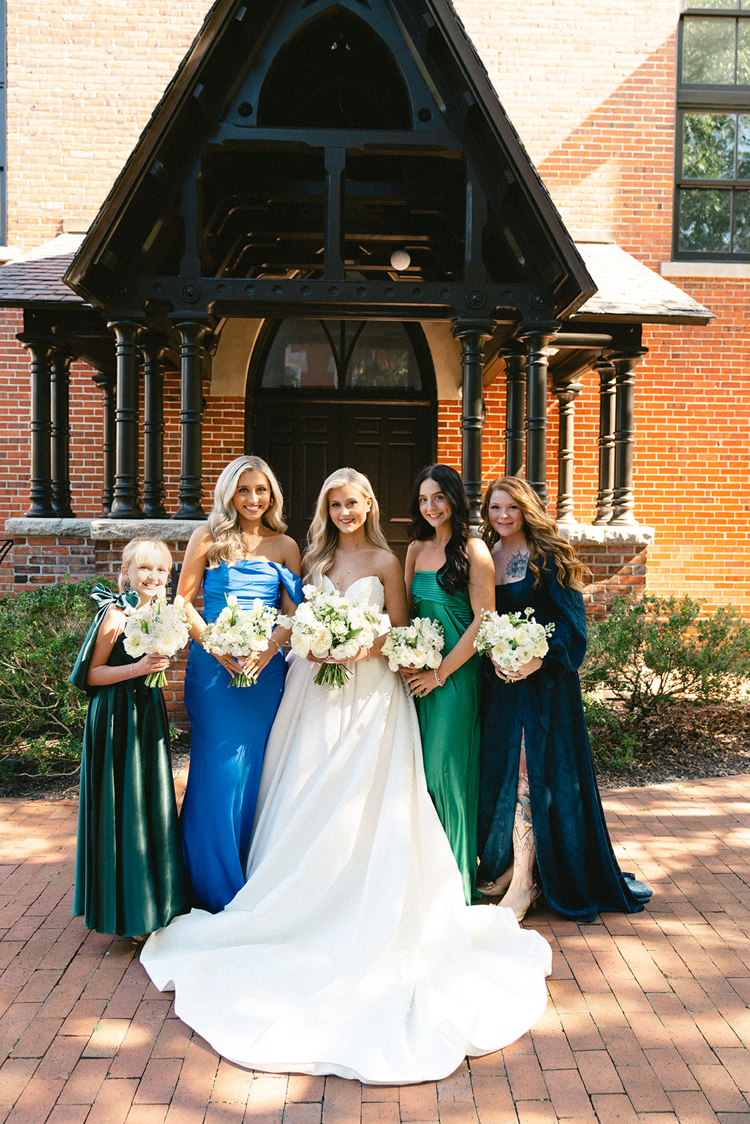 Five women in formal dresses stand together outdoors, holding bouquets, in front of a brick building with a black wooden porch.