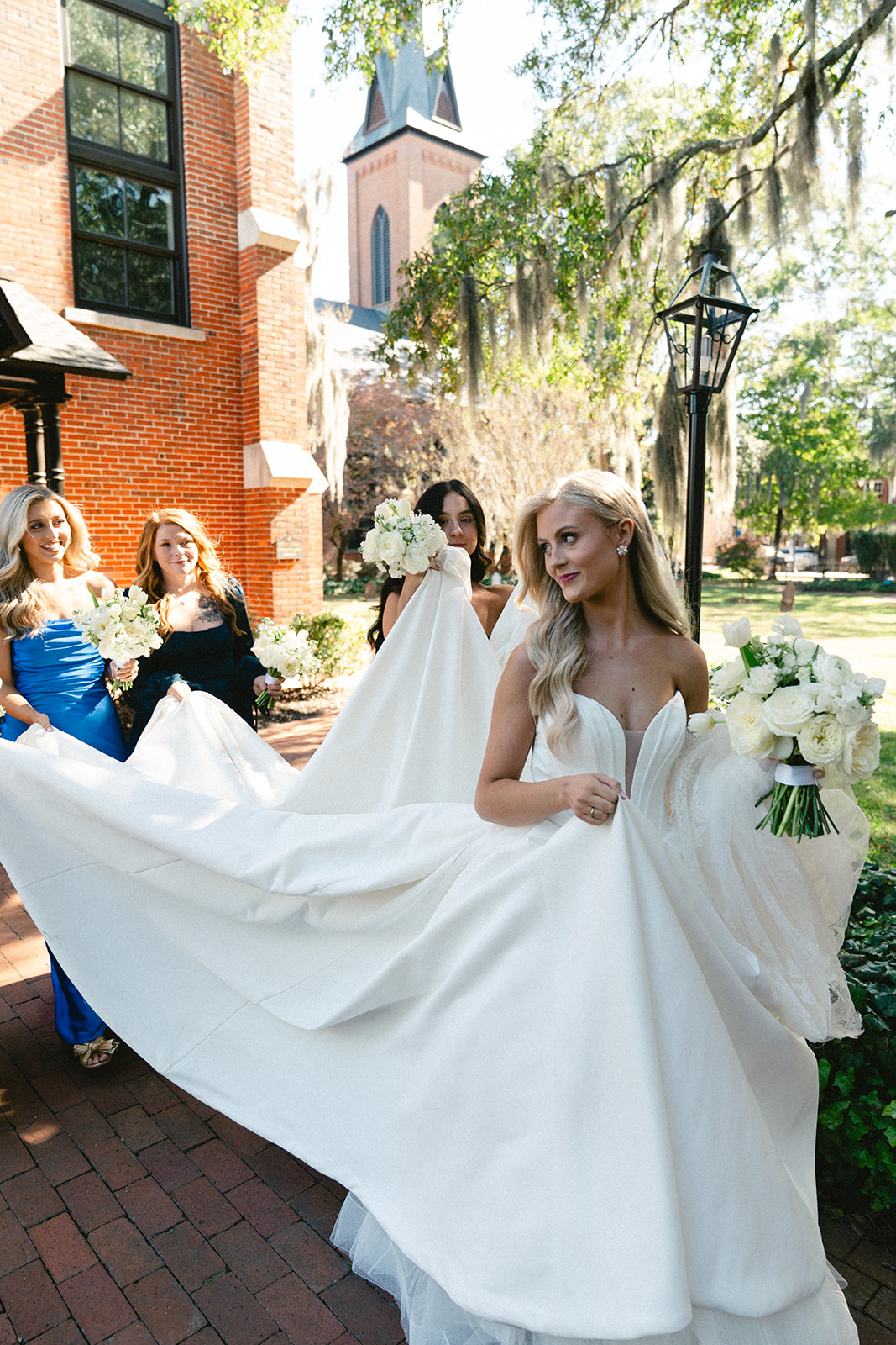 Five women in formal dresses stand together outdoors, holding bouquets, in front of a brick building with a black wooden porch.