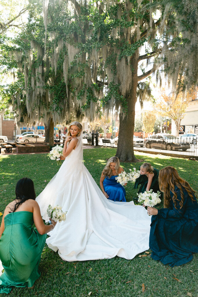 Five women in formal dresses stand together outdoors, holding bouquets, in front of a brick building with a black wooden porch.