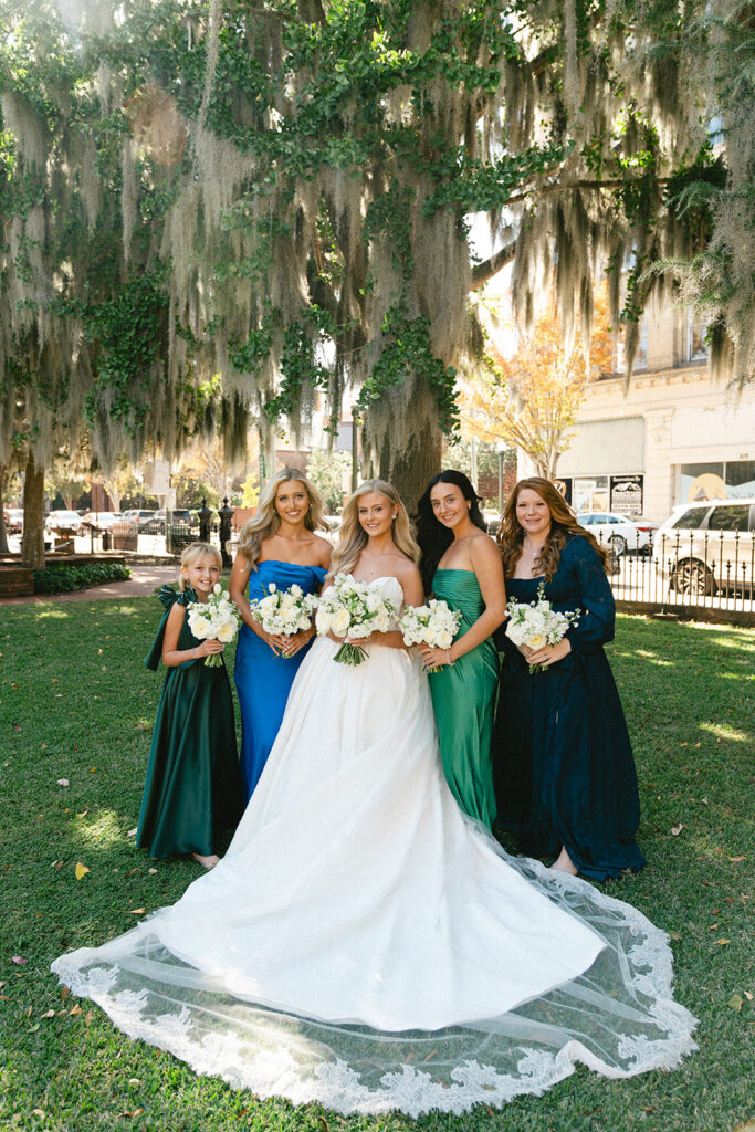 Five women in formal dresses stand together outdoors, holding bouquets, in front of a brick building with a black wooden porch.