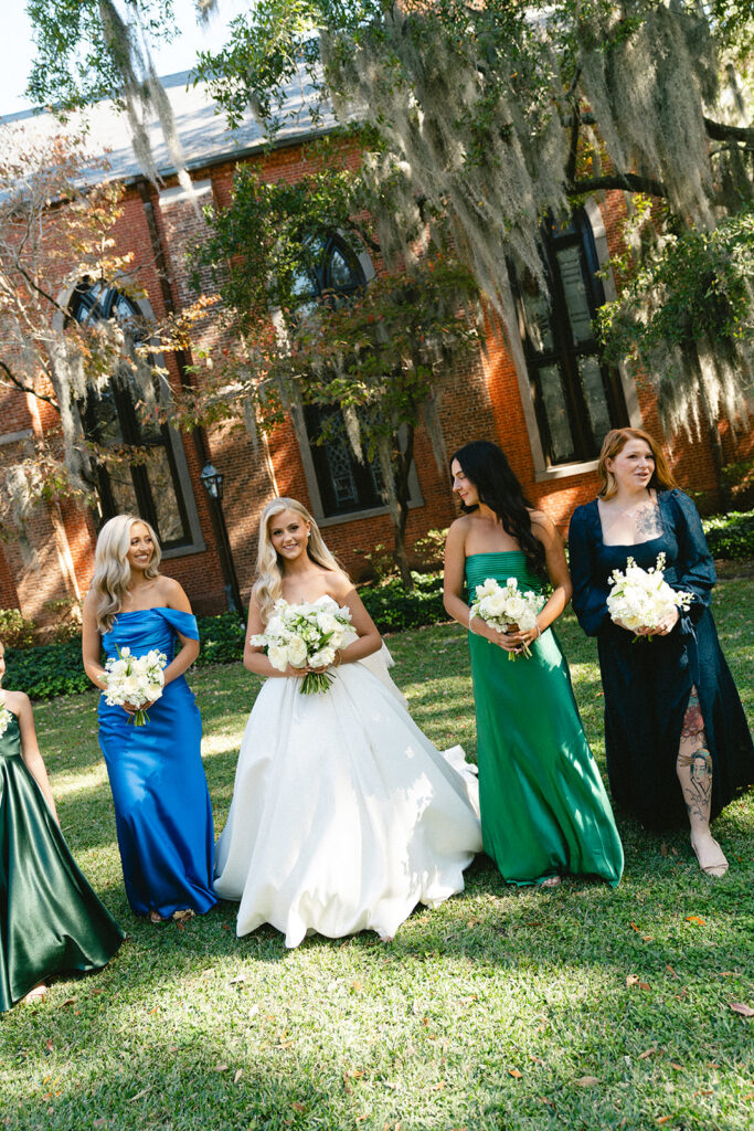 Five women in formal dresses stand together outdoors, holding bouquets, in front of a brick building with a black wooden porch.