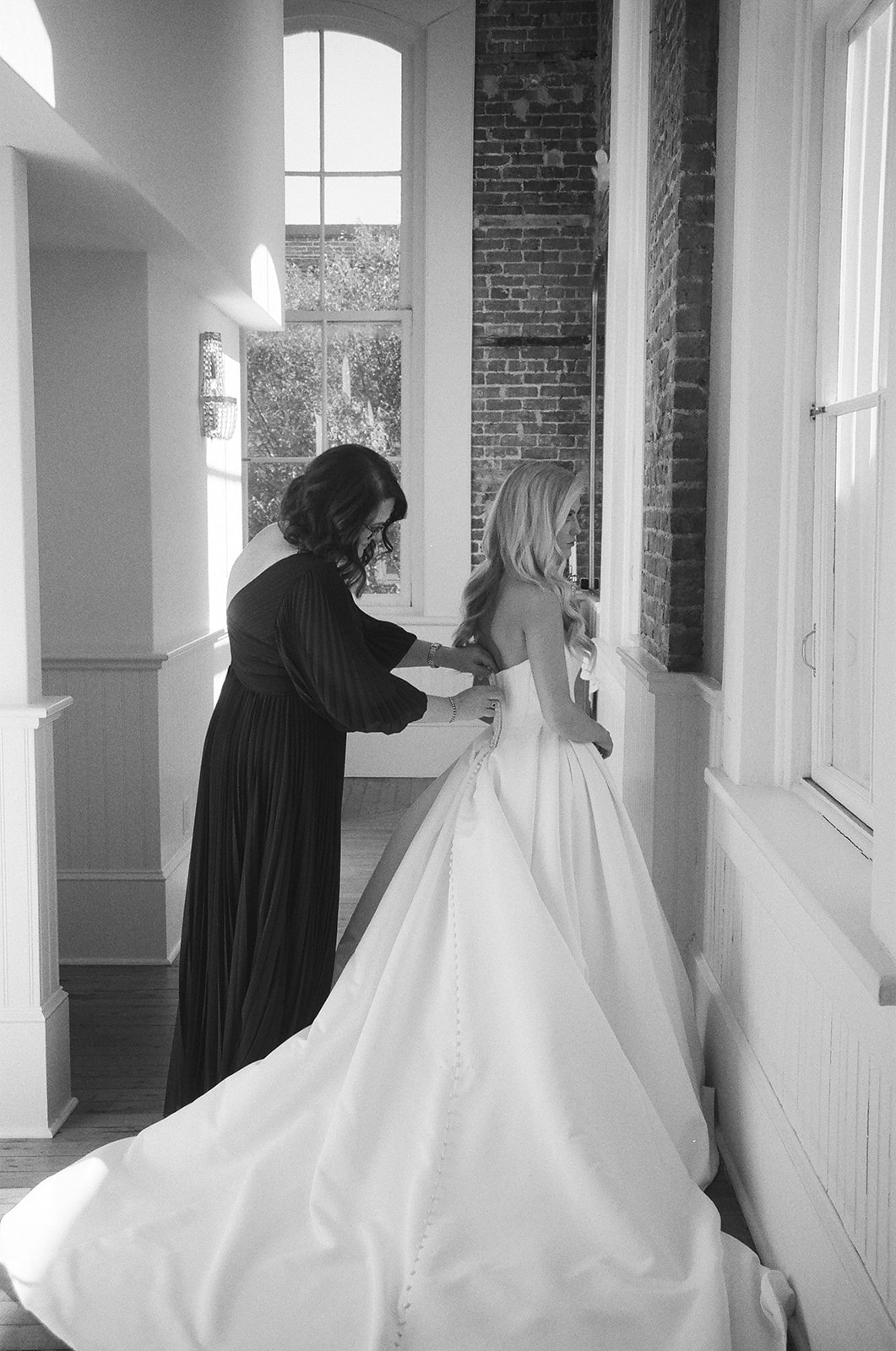 A woman in a dark dress helps another woman adjust her strapless wedding gown in a sunlit hallway with large windows for a wedding at Stanly Hall Ballroom