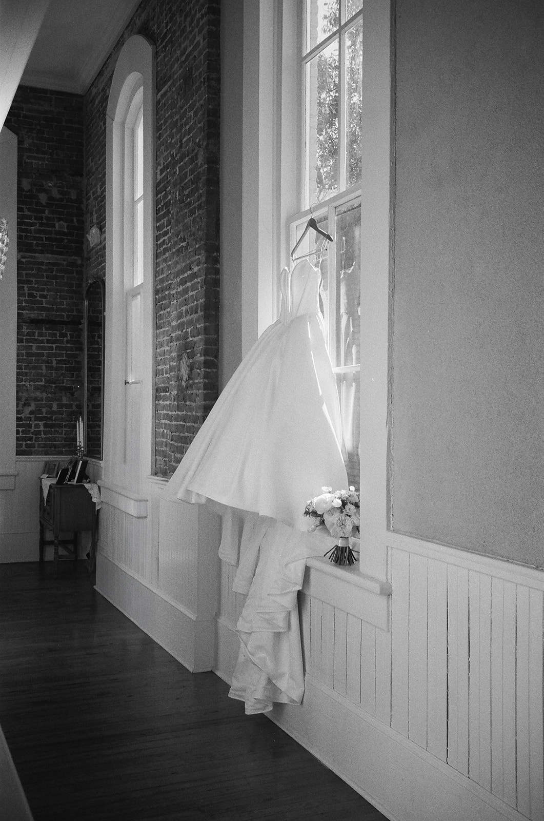 A wedding dress hangs on a window frame in a bright room with brick and white-paneled walls; a bouquet rests on the windowsill.