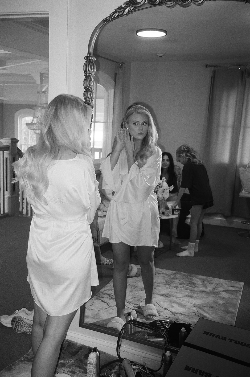 A woman in a white robe looks at herself in a large ornate mirror while touching her hair; others are seated in the background of the room for a wedding at Stanly Hall Ballroom