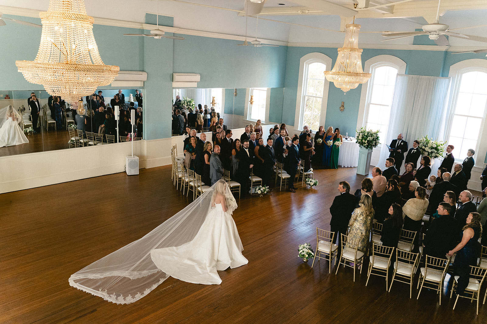 A decorated wedding ceremony setup with gold chairs, floral arrangements, and large windows in a light blue room with wooden floors at Stanly Hall Ballroom