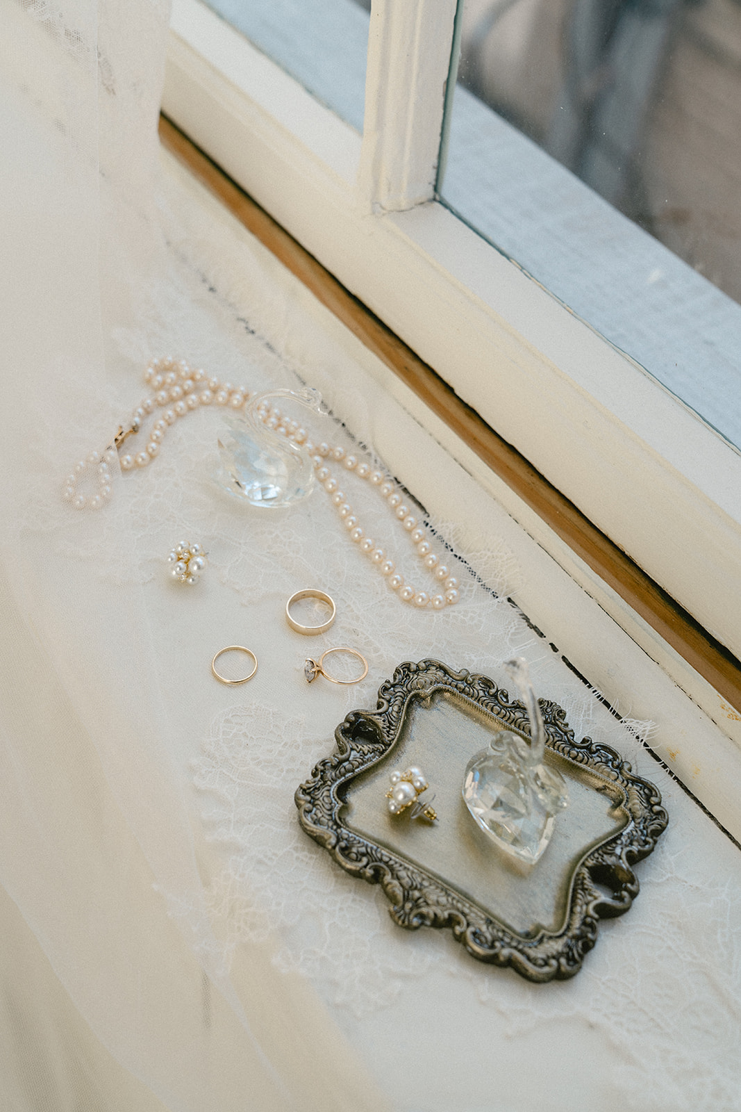 Wedding invitation flat lay with lace veil, jewelry, pearl necklace, rings on a tray, white flowers, and a tulip on a wooden surface.