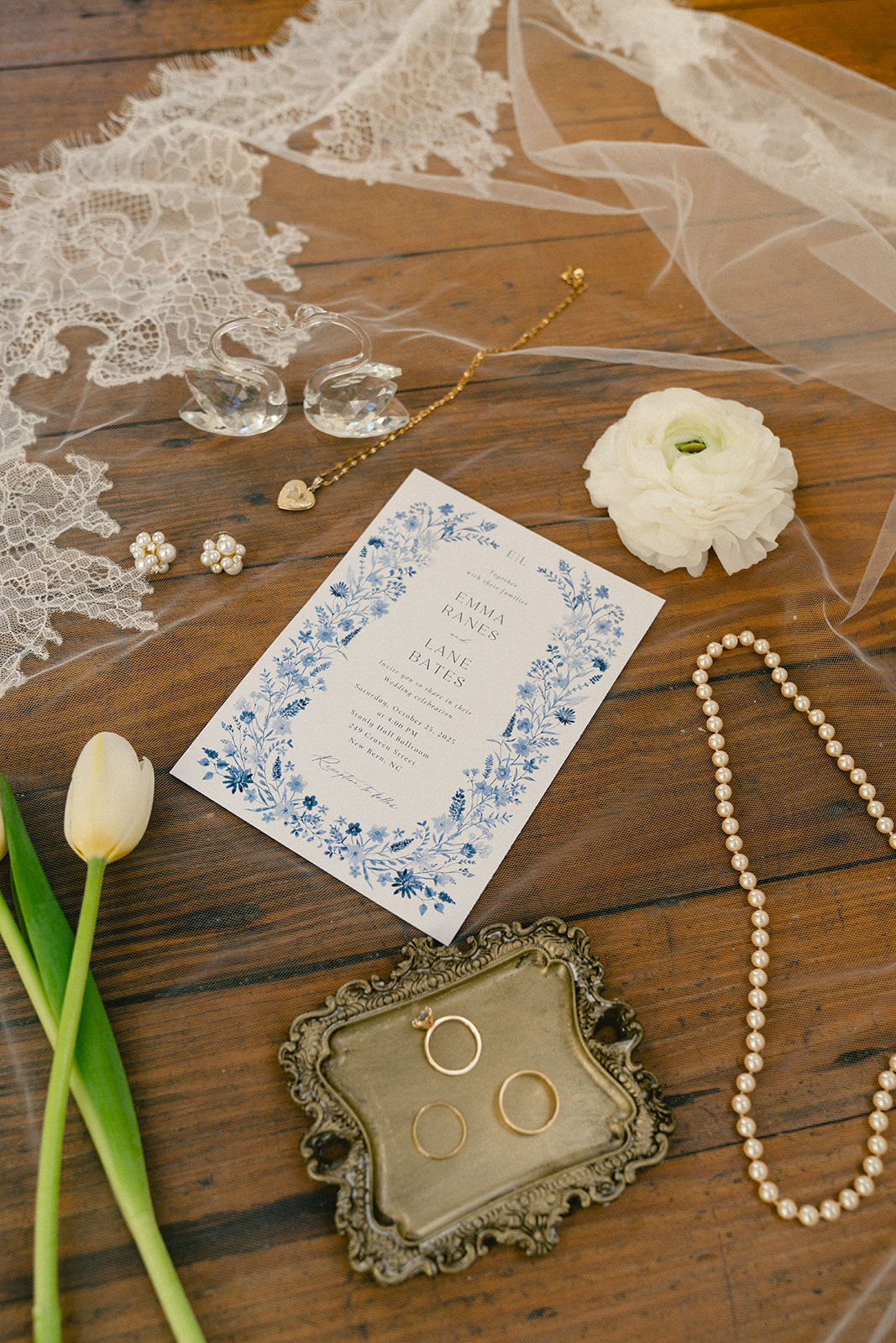 Wedding invitation flat lay with lace veil, jewelry, pearl necklace, rings on a tray, white flowers, and a tulip on a wooden surface.