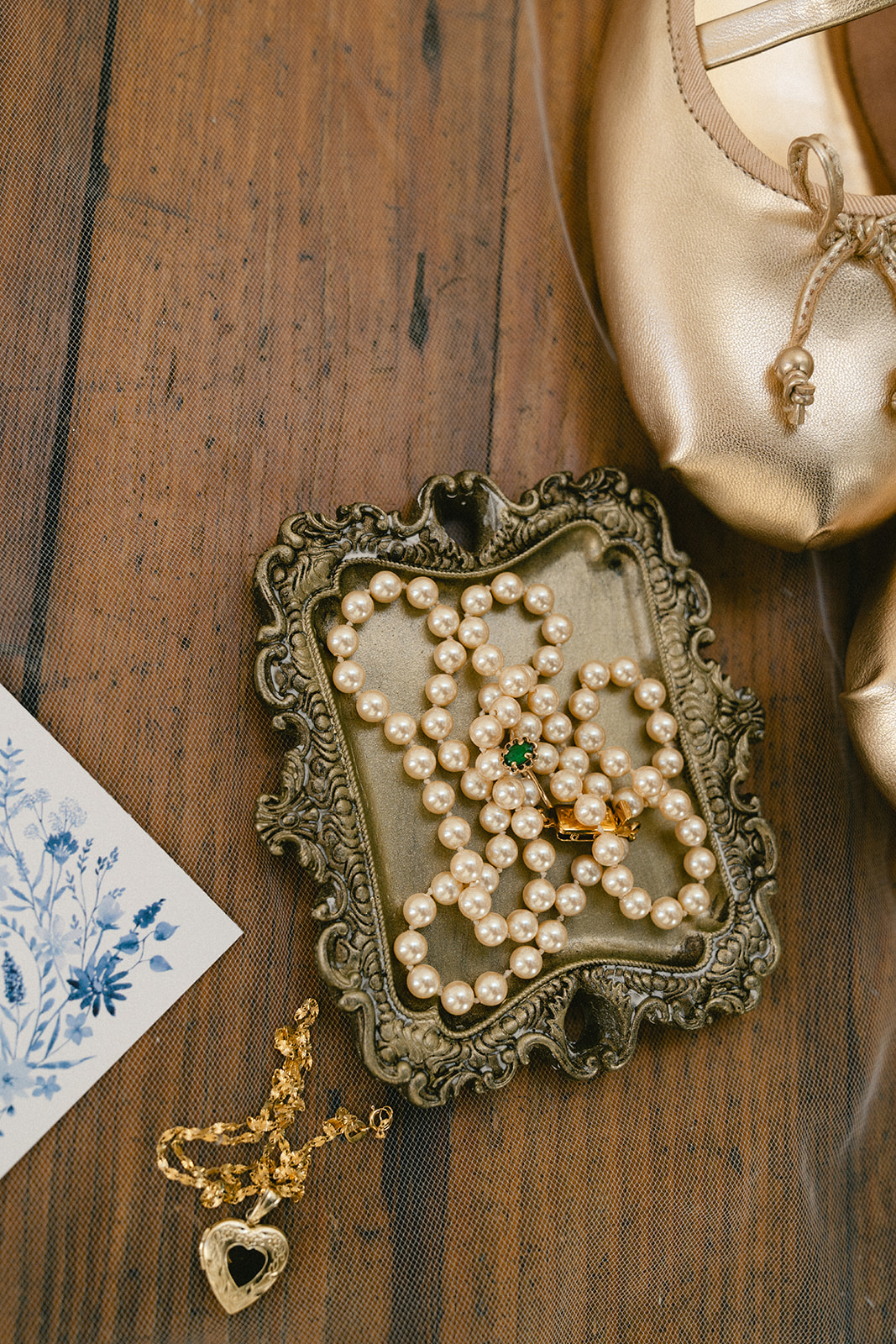 Wedding invitation flat lay with lace veil, jewelry, pearl necklace, rings on a tray, white flowers, and a tulip on a wooden surface.