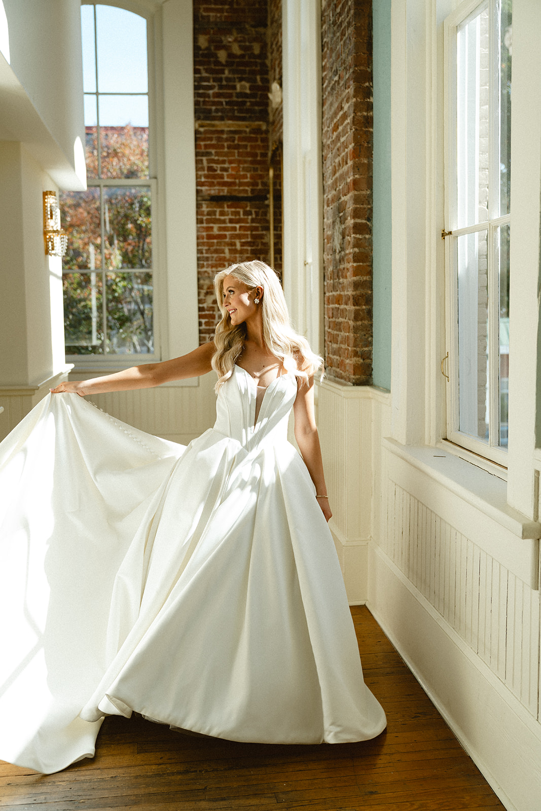 A woman in a white strapless wedding gown stands indoors by large windows and exposed brick walls, with sunlight streaming in at Stanly Hall Ballroom