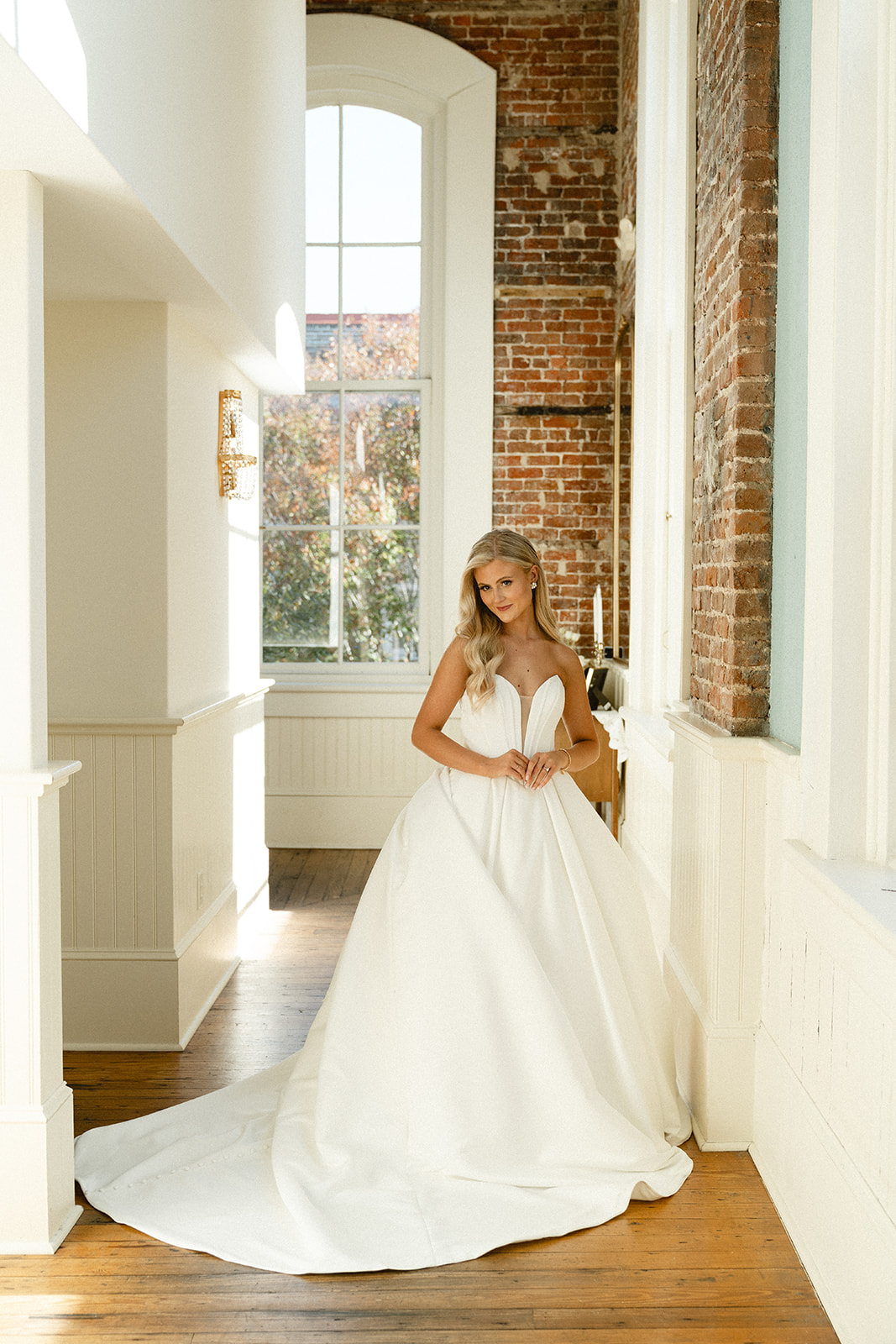 A woman in a white strapless wedding gown stands indoors by large windows and exposed brick walls, with sunlight streaming in at Stanly Hall Ballroom