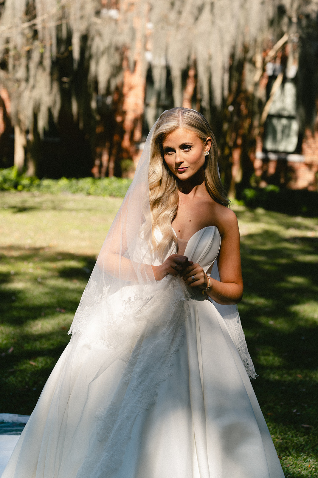 A bride in a strapless white wedding dress and veil stands outdoors, partially in sunlight and shadow, looking over her shoulder.