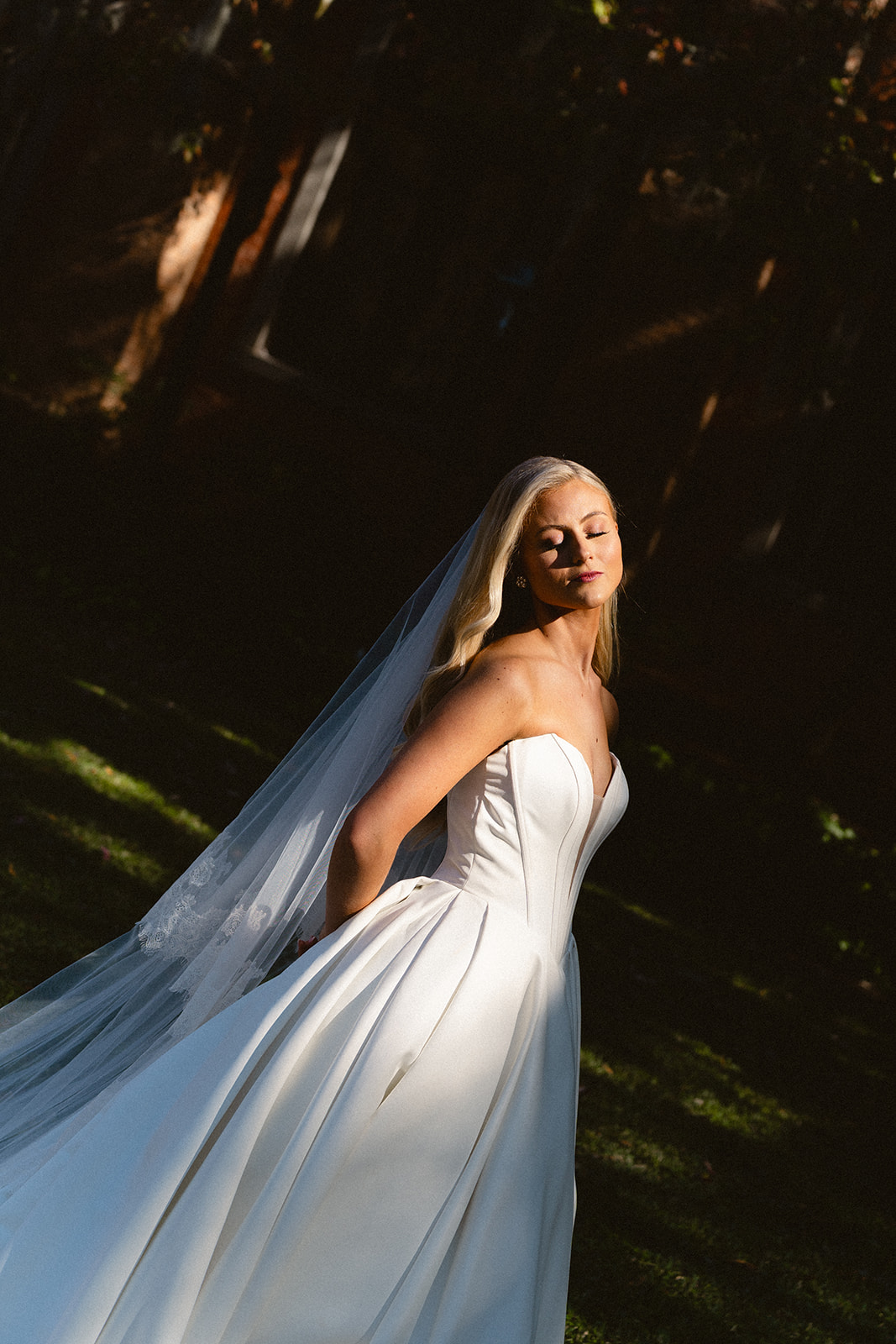 A bride in a strapless white wedding dress and veil stands outdoors, partially in sunlight and shadow, looking over her shoulder.