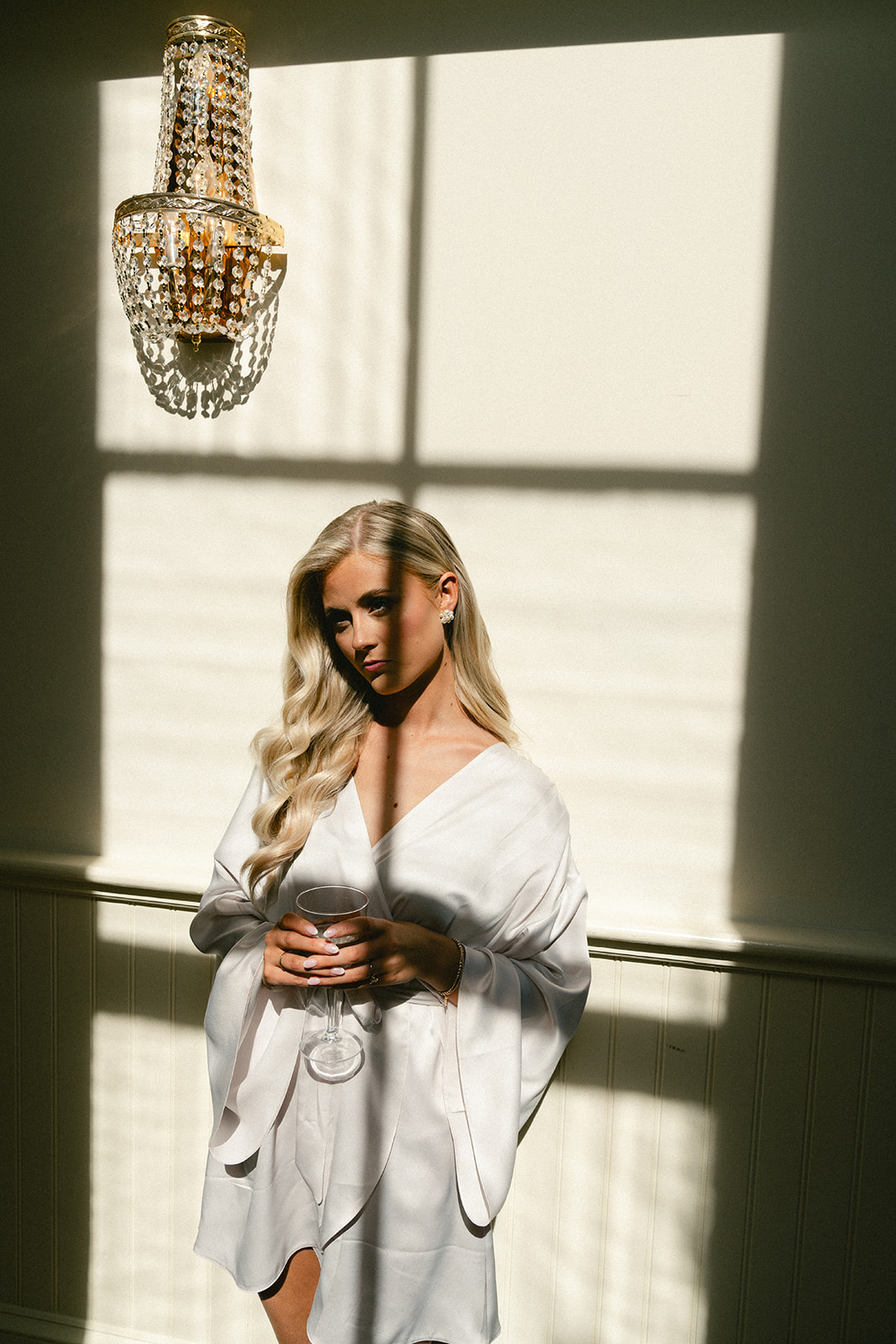 A woman in a white robe stands in sunlight, holding a cup, with shadows from a window grid and a chandelier visible on the wall behind her.