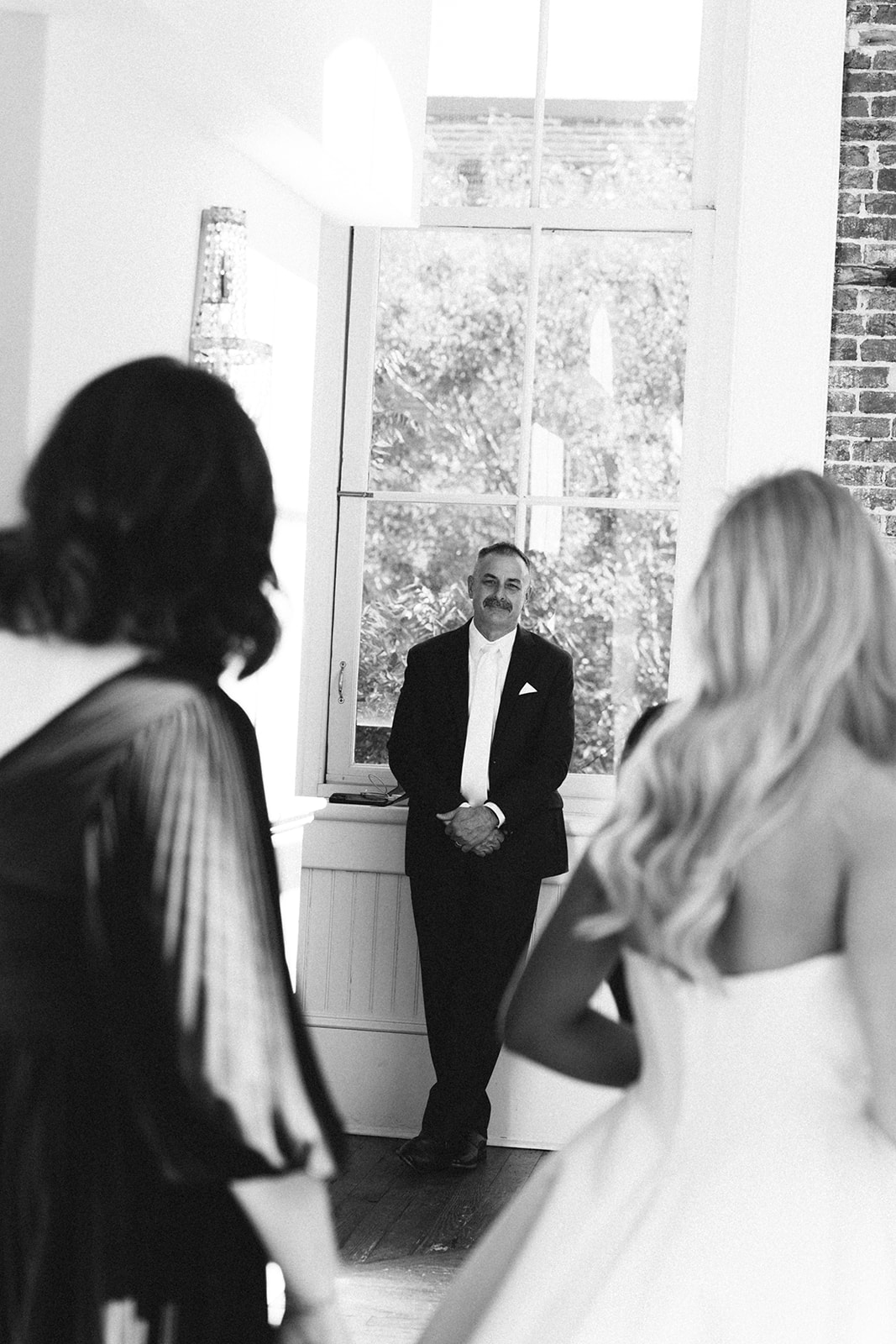 A man in a suit stands by a large window, facing two women in formal dresses, one in a wedding gown, in a brightly lit room.