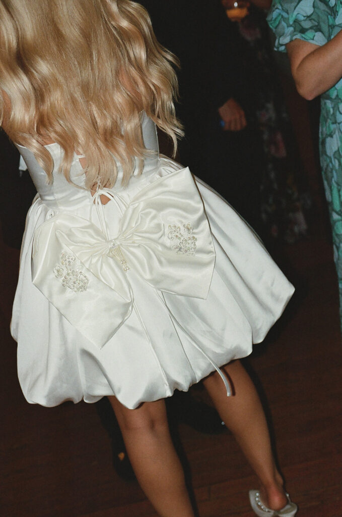 Person wearing a white dress with a large bow on the back, standing indoors on a wooden floor.