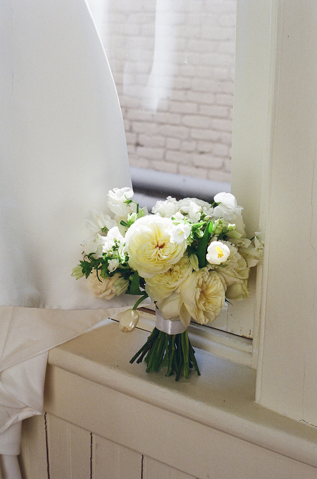 A glass vase with white flowers sits on a windowsill beside a partially open window and white curtains.