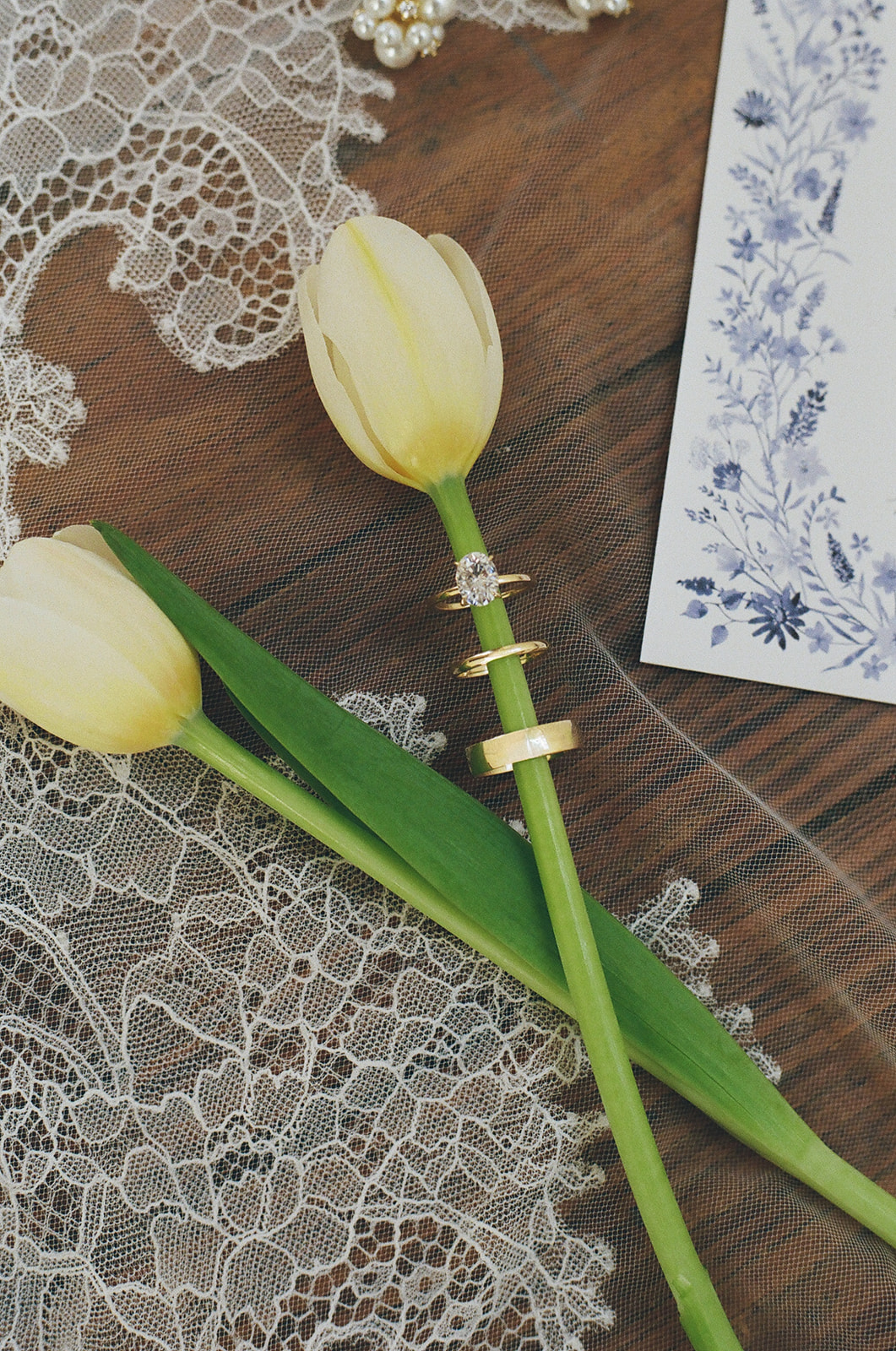 Two yellow tulips with rings on their stems rest on lace fabric next to a floral-patterned card on a wooden surface.