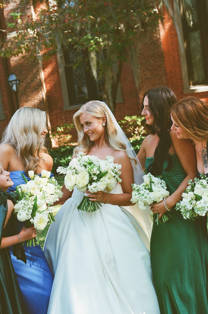 A bride in a white dress stands outdoors with four bridesmaids in colorful dresses, all holding white flower bouquets and smiling at each other.