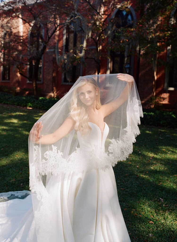 A bride in a white wedding dress holds up a lace-trimmed veil while standing on grass in front of a brick building.