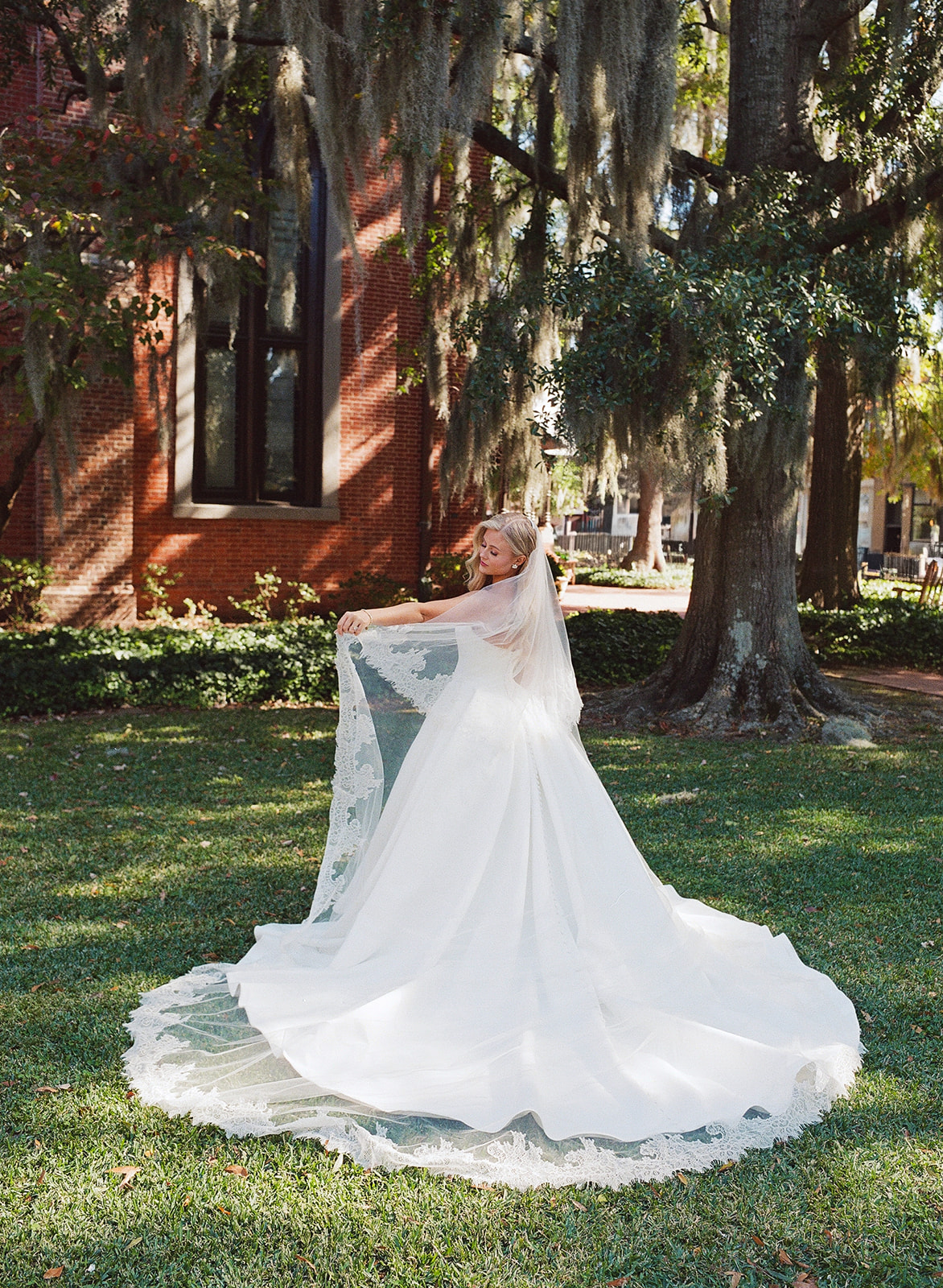 A bride in a white wedding dress holds up a lace-trimmed veil while standing on grass in front of a brick building.
