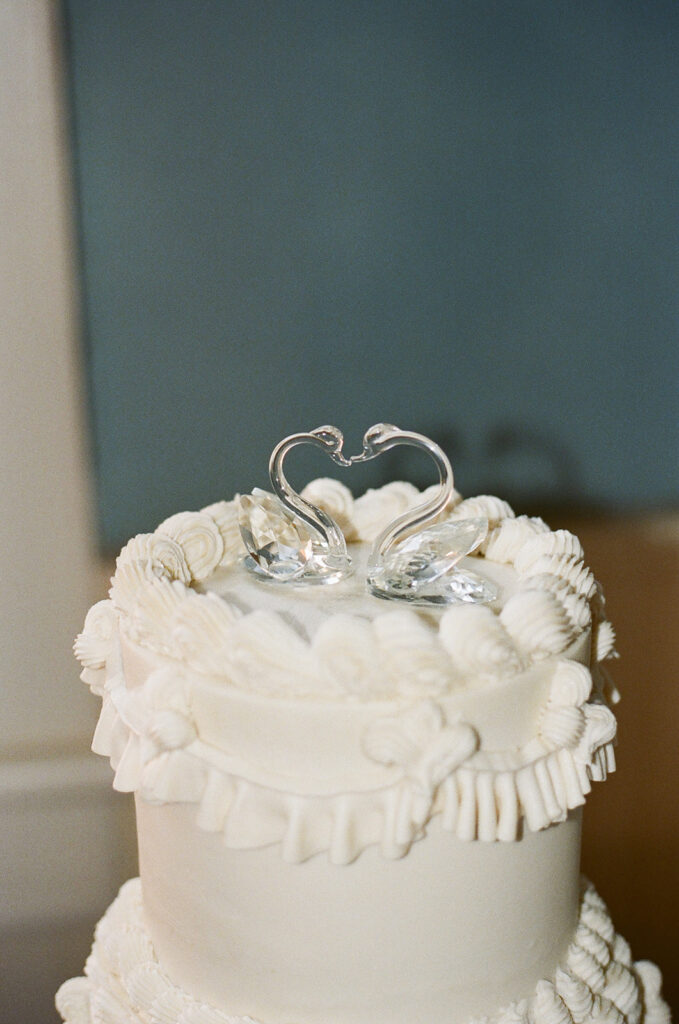 White wedding cake with decorative icing and a clear glass topper shaped like two swans forming a heart.