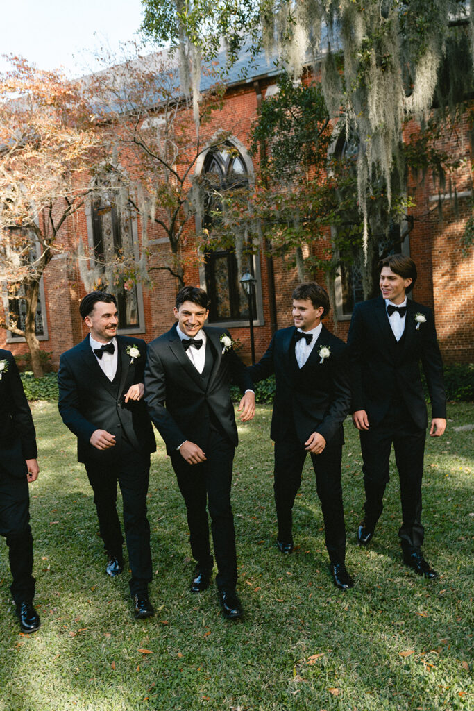 Five men in black tuxedos walk together on grass outside a brick building with arched windows and autumn trees in the background.