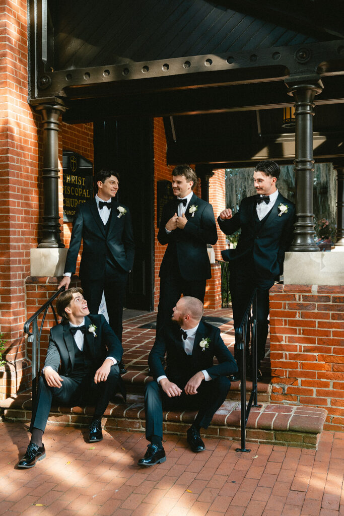 Five men dressed in black tuxedos with white boutonnieres stand and sit on steps outside a brick building, engaged in conversation.
