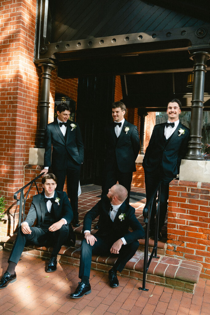 Five men dressed in black tuxedos with white boutonnieres stand and sit on steps outside a brick building, engaged in conversation.
