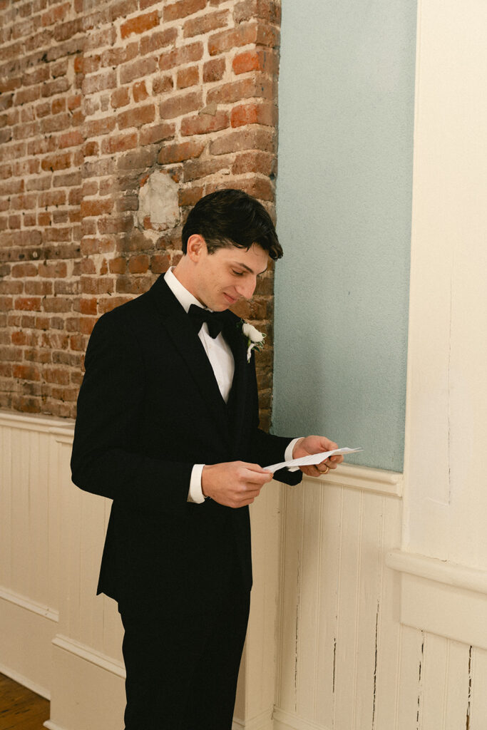 A man in a black tuxedo reads a piece of paper while standing indoors near a brick and paneled wall.