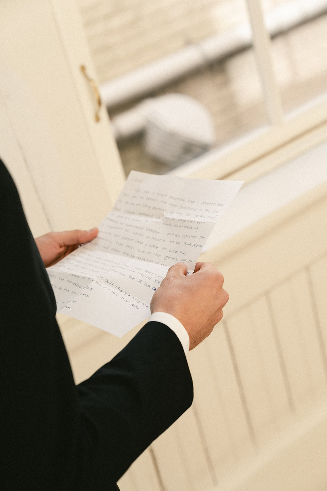 A man in a black tuxedo reads a piece of paper while standing indoors near a brick and paneled wall.