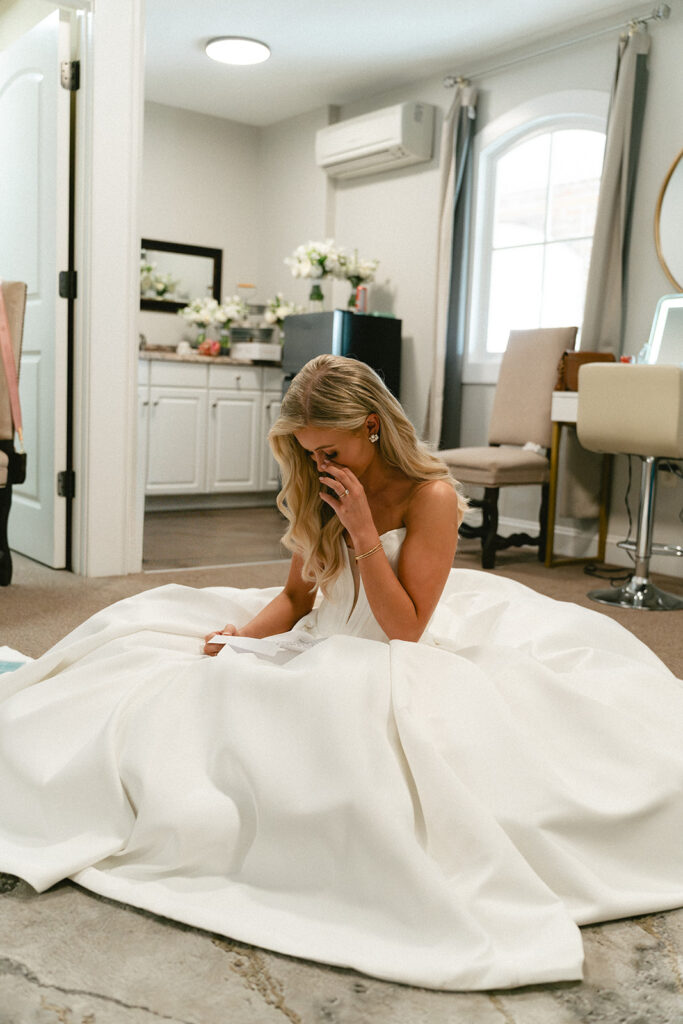 A woman in a white wedding dress sits on the floor of a dressing room, wiping her eyes with her hand.