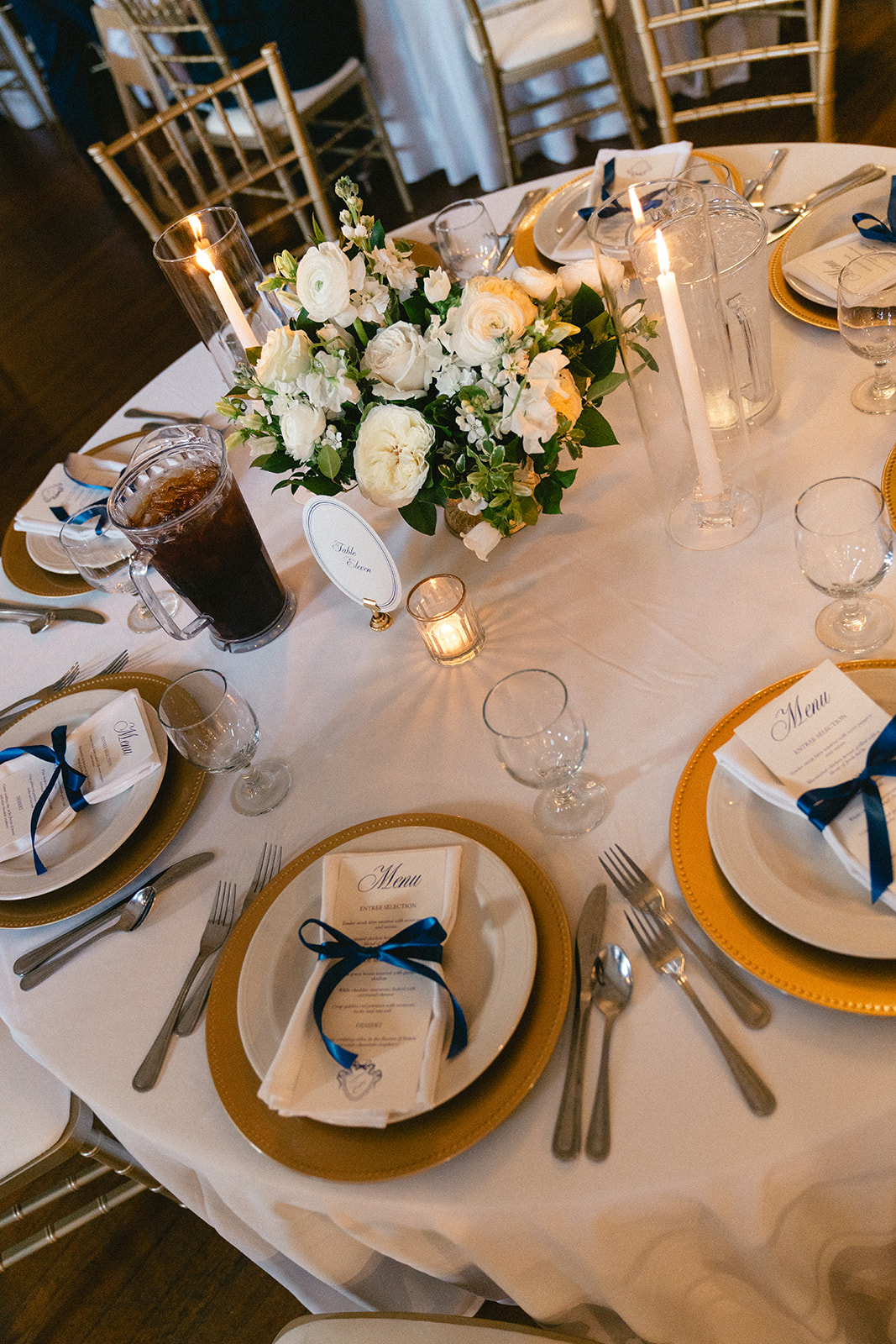 Round table set for an event with gold plates, white napkins tied with blue ribbon, floral centerpiece, candles, and glassware.