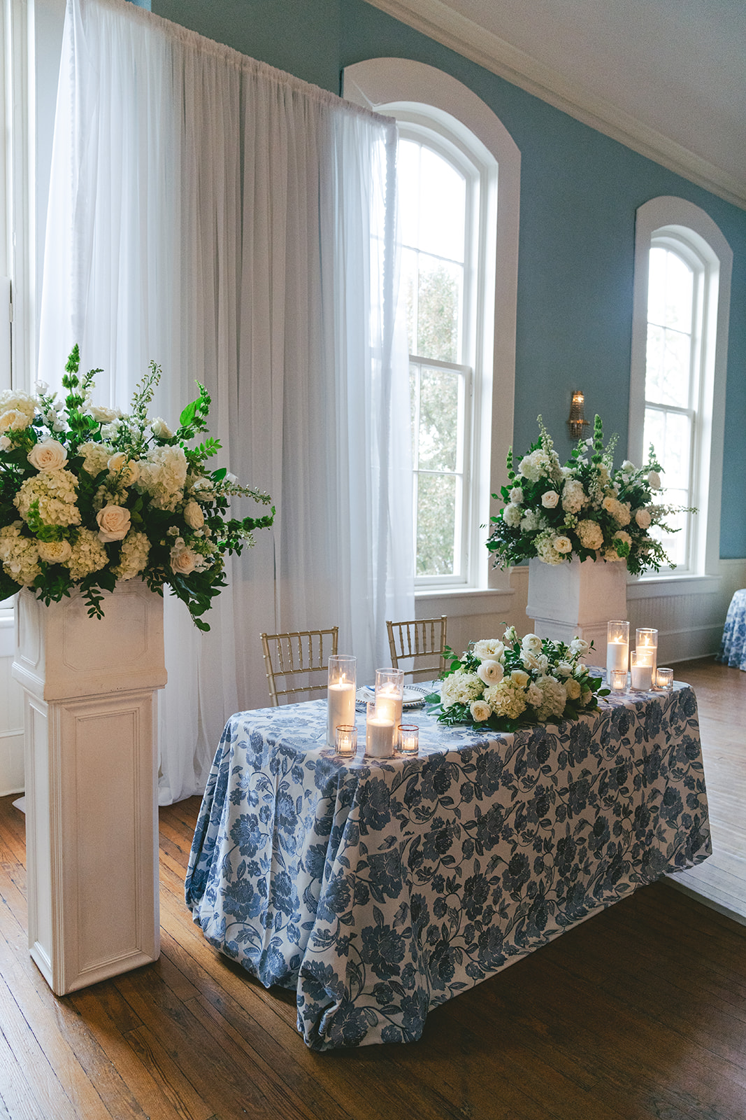 A table with a blue and white floral tablecloth is decorated with white flower arrangements and candles, set in front of tall windows with sheer curtains.