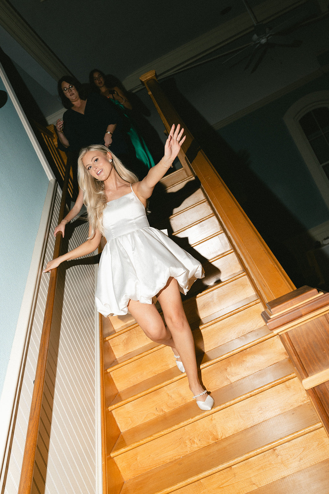 Three women walk down a wooden staircase, with the woman in front wearing a short white dress and smiling at the camera at Stanly Hall Ballroom