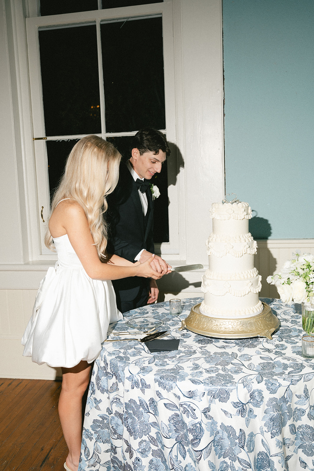 A couple dressed formally cuts a white tiered wedding cake together on a round table with a blue floral tablecloth.