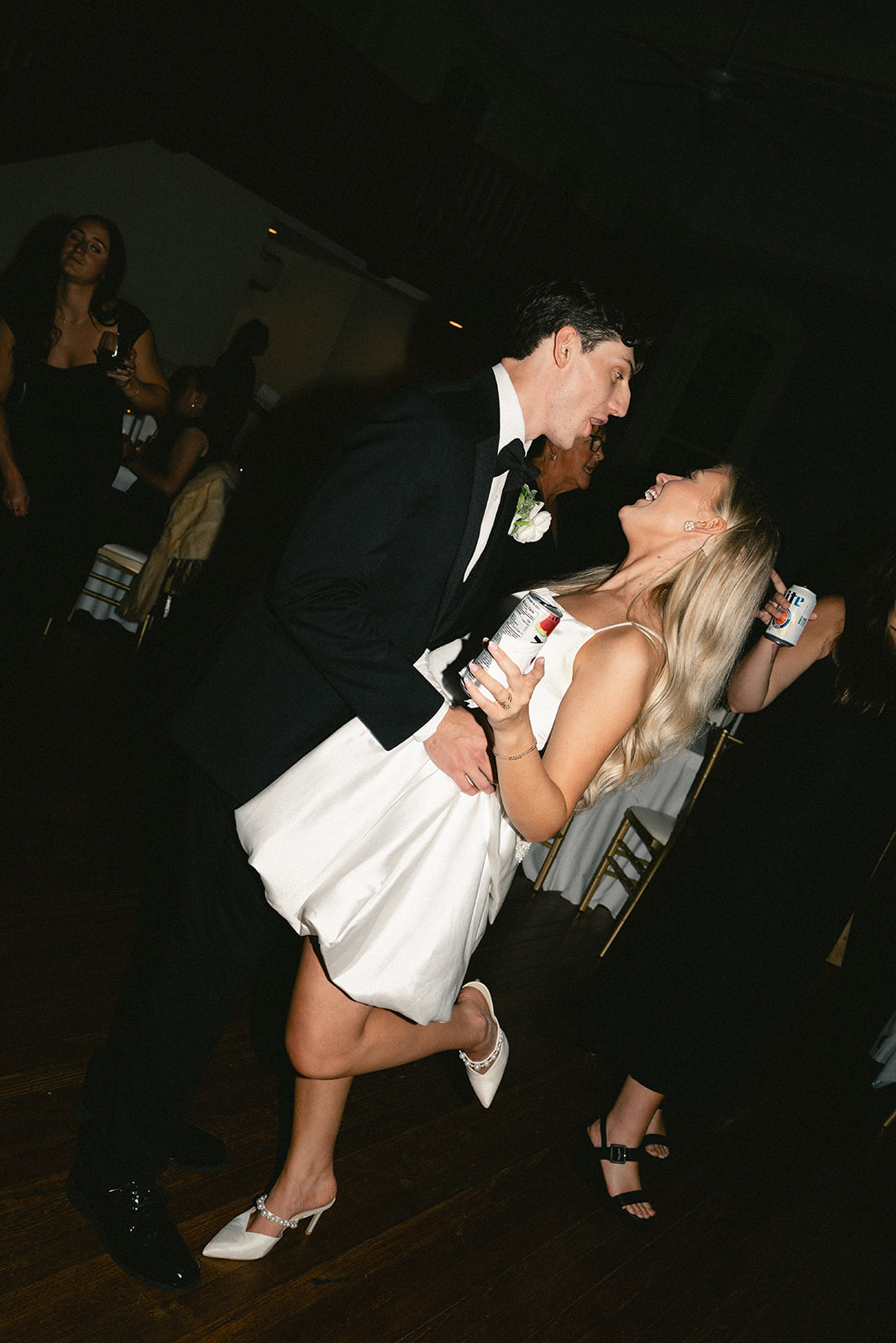 A man in a black suit and a woman in a white dress are dancing together at an indoor event, surrounded by other people.