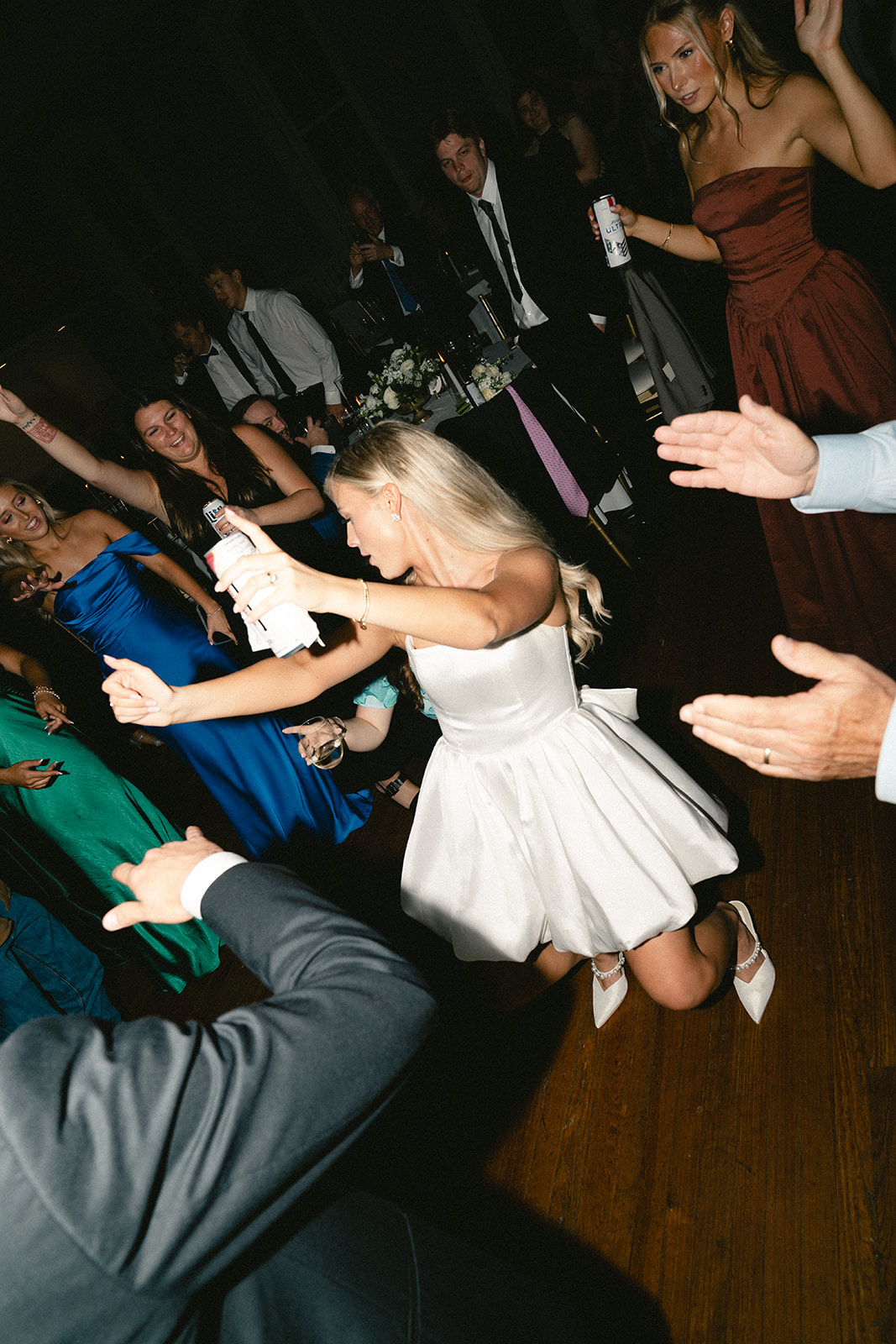 A woman in a white dress dances enthusiastically on the floor at a lively wedding surrounded by people in formal attire.