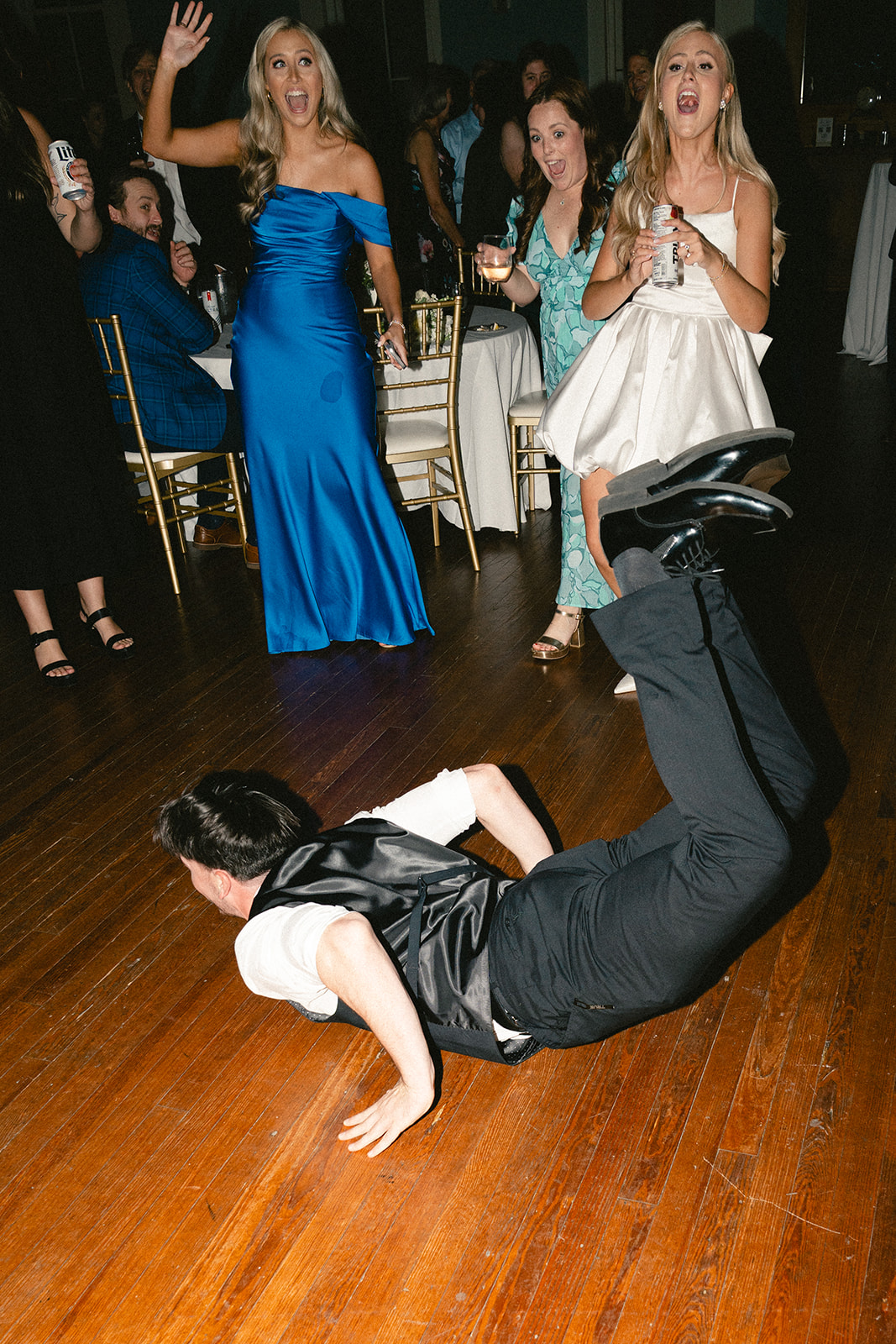 Man dances enthusiastically on the floor at a lively wedding surrounded by people in formal attire.