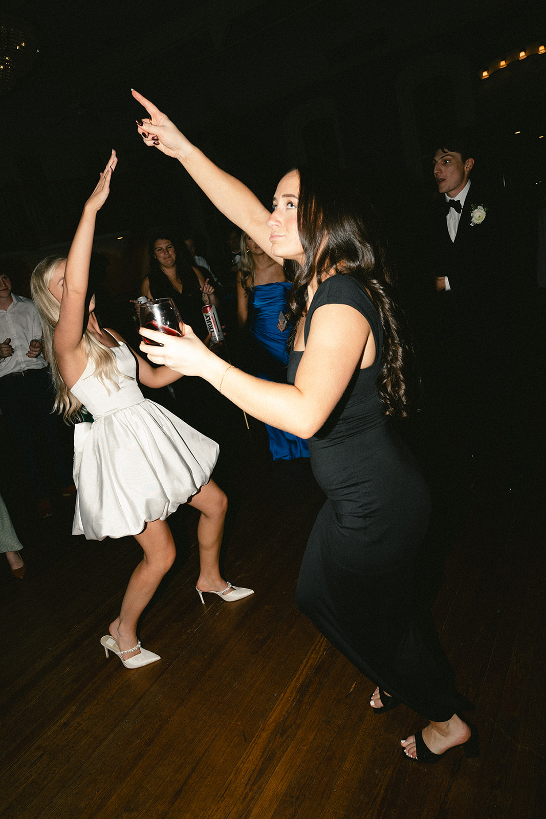 A woman in a white dress dances enthusiastically on the floor at a lively wedding surrounded by people in formal attire.