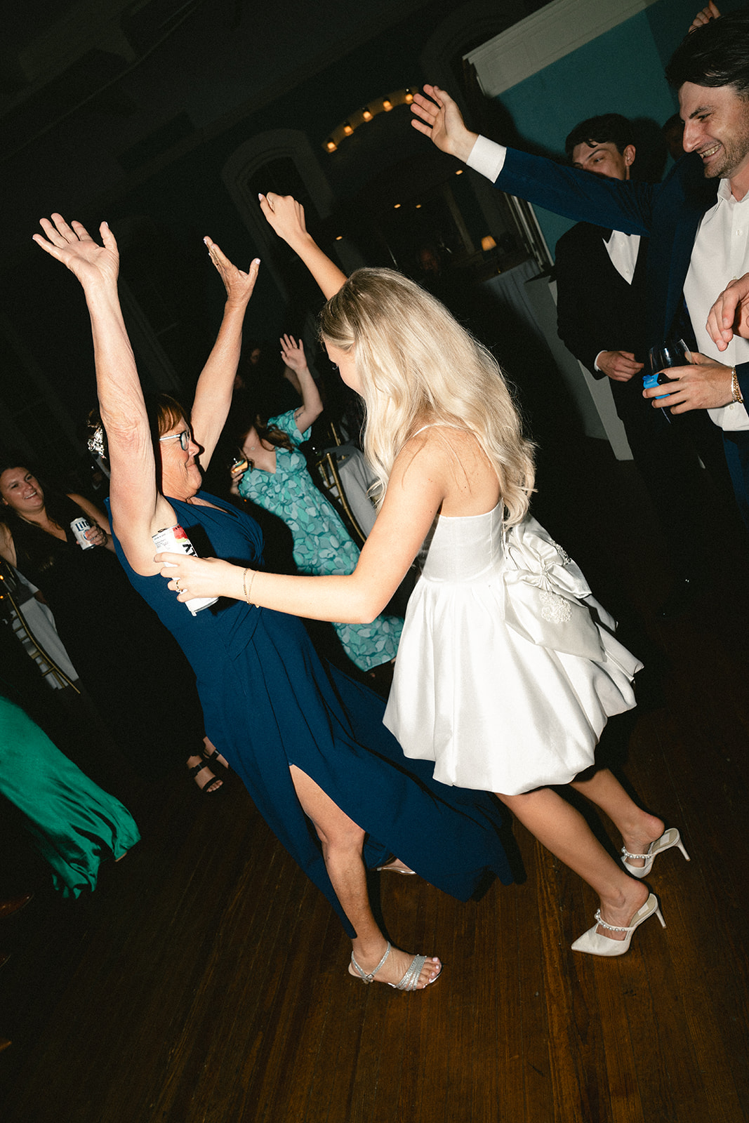 A woman in a white dress dances enthusiastically on the floor at a lively wedding surrounded by people in formal attire.