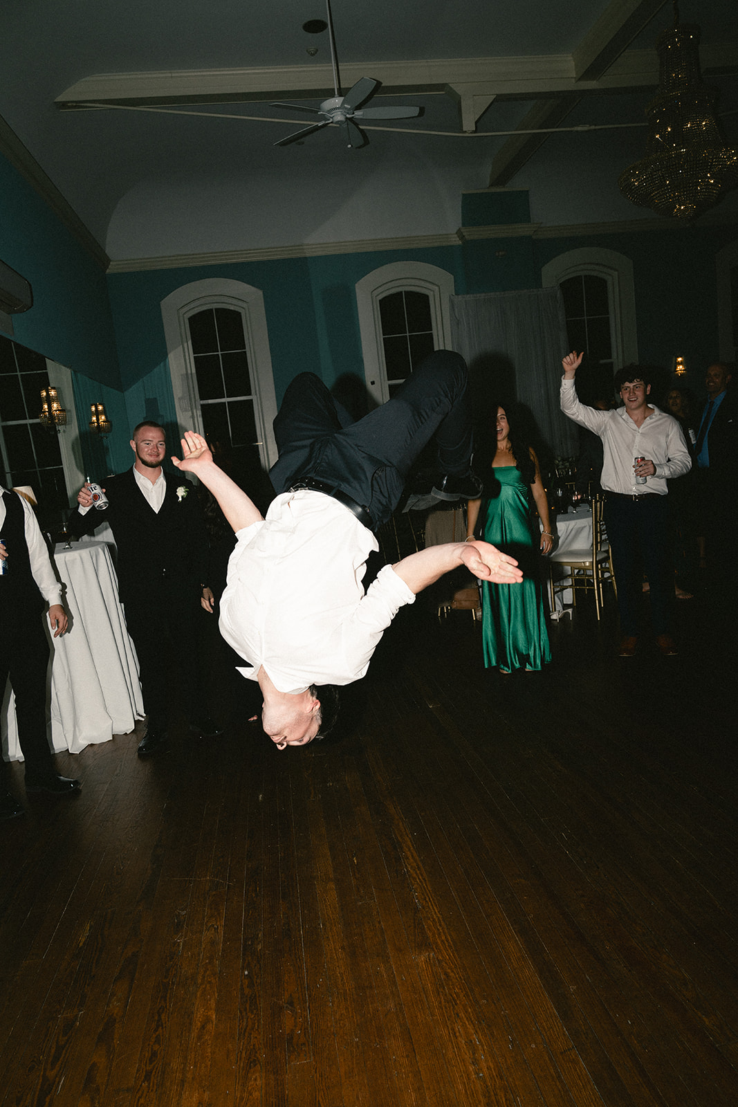 Man dances enthusiastically on the floor at a lively wedding surrounded by people in formal attire.